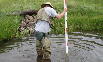 Field crew member standing in a wadeable stream, using a leveling rod to measure stream depth.