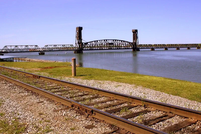 Railroad tracks alongside a large river, with a bridge in the distance.